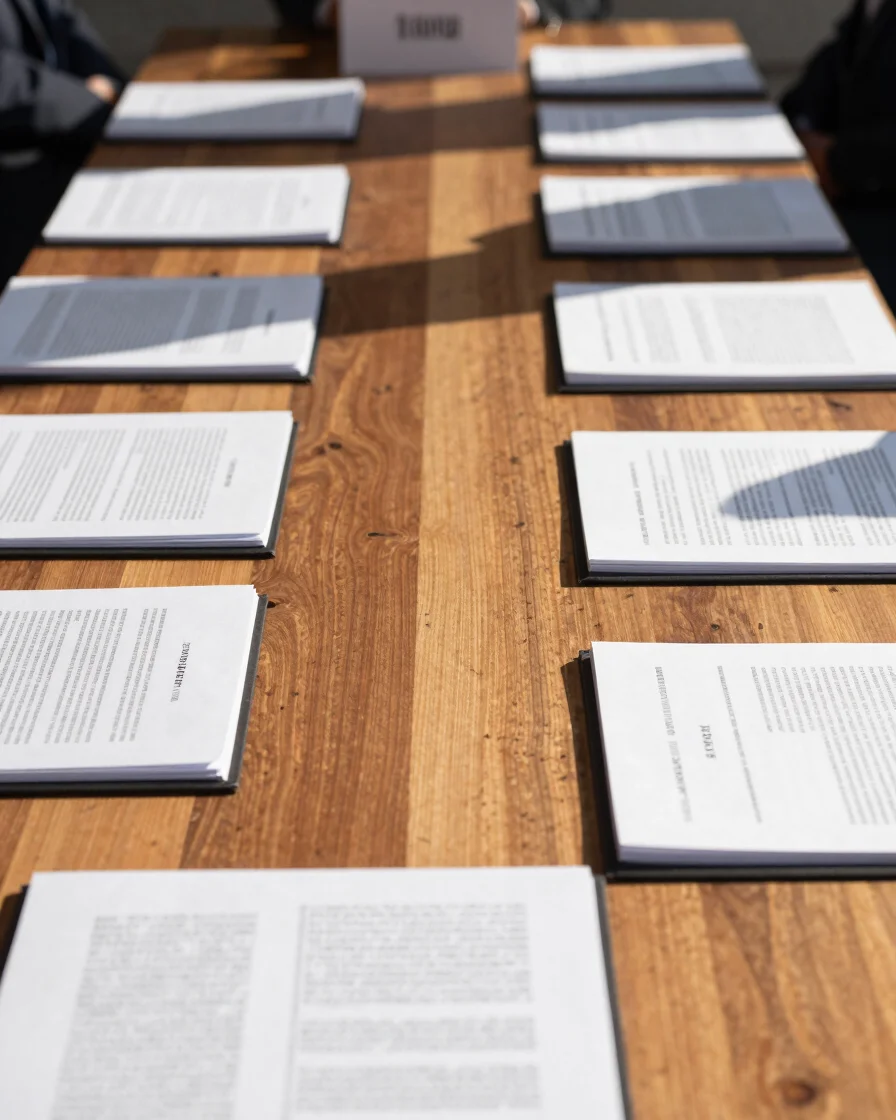 Journalistic wide shot of a field press briefing with document folders on a wooden table under crisp natural sunlight